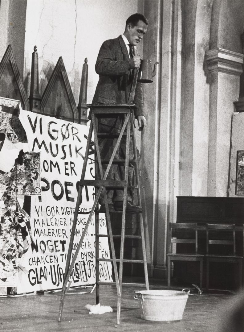 Black and white photograph of a man in suit and tie holding a tea kettle while standing on a wooden ladder. At the base of the ladder sits a metal tub. Behind the ladder sits a large sign with painted words in Danish.
