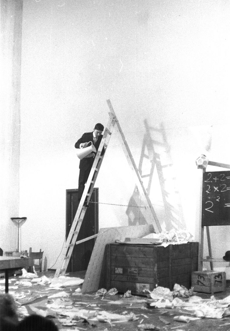 Black and white photograph of a man in a suit and tie holding a pitcher while standing on an over ten-foot-tall ladder. Loose pages are strewn about the floor surrounding the ladder, while the high-ceilinged wall behind the ladder is blank.