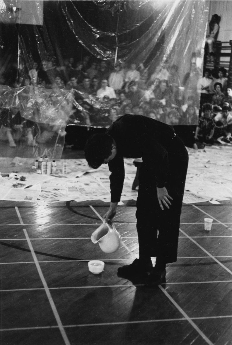 Black and white photograph of a man bending over to pour water from a ceramic pitcher to a small bowl sitting on the floor below. In the background, an audience views the performance from behind clear plastic sheeting.
