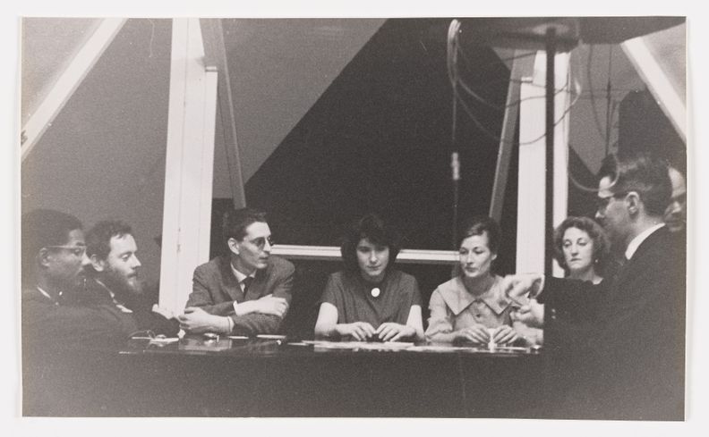 Black-and-white landscape-oriented photograph of four men and three women sitting indoors in an arc around a table. Most are looking to the man seated furthest right whose hands are in motion.
