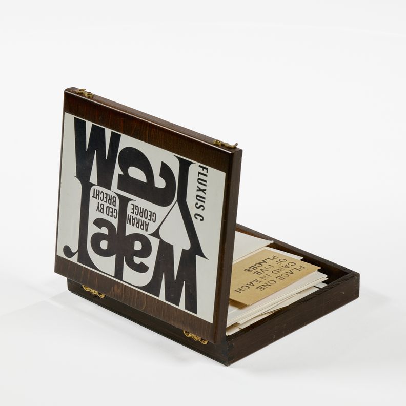 Top view of wooden box dominated by a white label with large black letters that read, Water Yam. Smaller text surrounding reads, FLUXUS C and ARRANGED BY GEORGE BRECHT. The hinged wooden box is open to reveal stacked cardstock. One card, brown unlike the other white cards, sticks out and has text, place one card in each of five places.