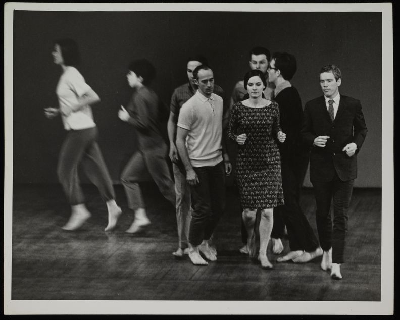 Horizontal black and white photograph of eight spot-lit people running barefoot in an otherwise empty room.