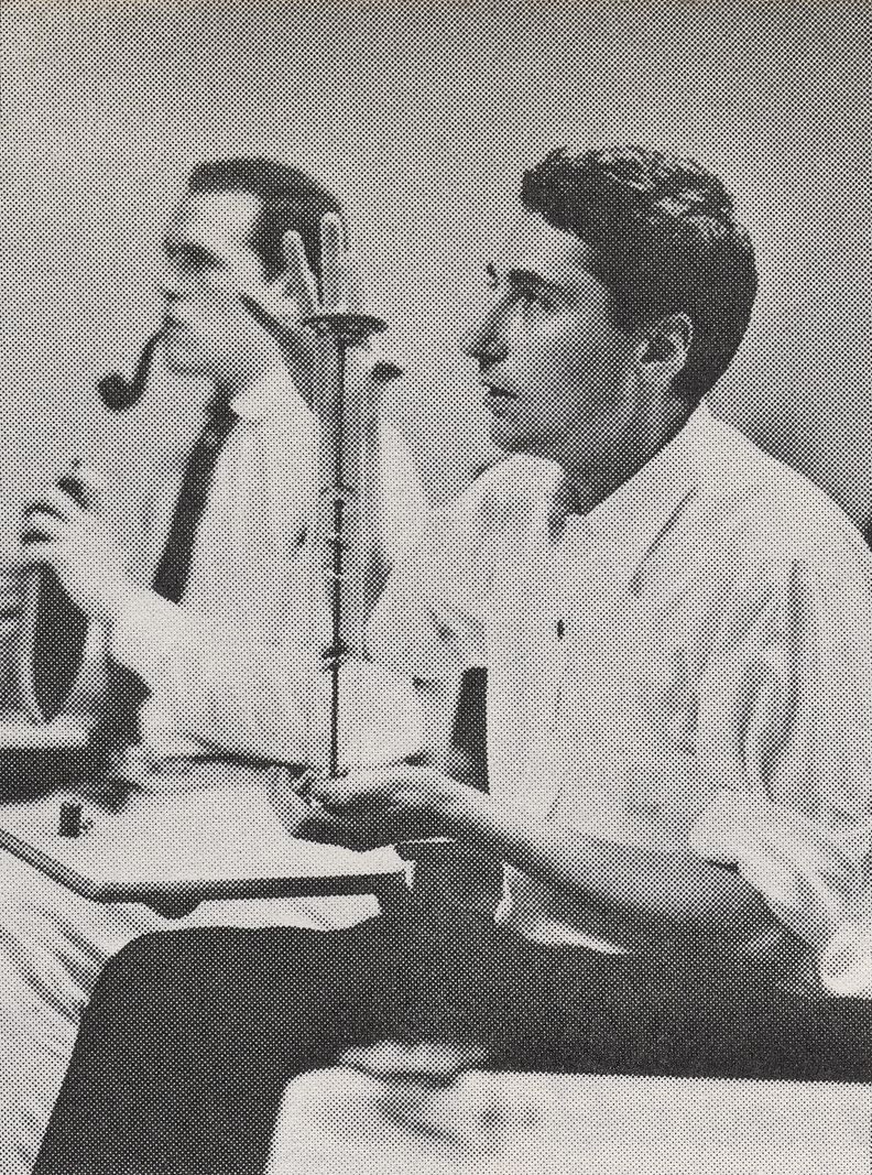 A vertical black and white photograph of two men looking toward a non visible speaker. Both men hold up different unidentifiable objects.