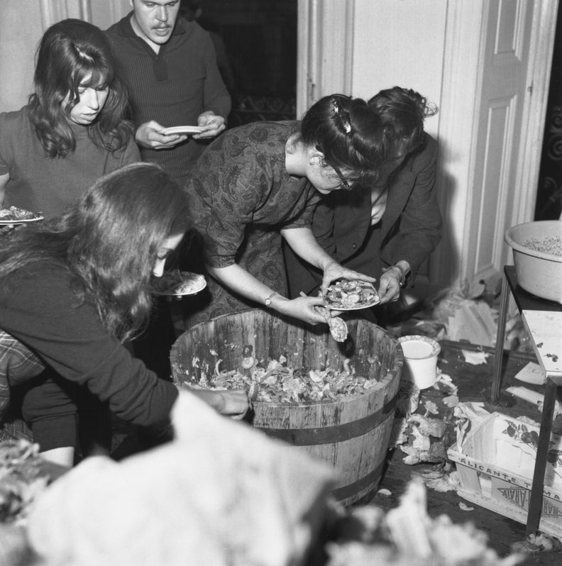 Square black and white photograph of five people indoors at top left filling plates from a large wooden bucket of salad at center.