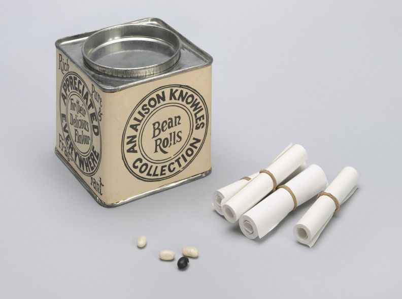 Color photograph of square cream painted metal tin with circular logo with text, An Alison Knowles Collection, on the outer rim and the words Bean Rolls displayed in the center. Four rubber-banded rolls of paper are displayed to the right of the tin and four beans, three white and one black, are displayed in front of tin.