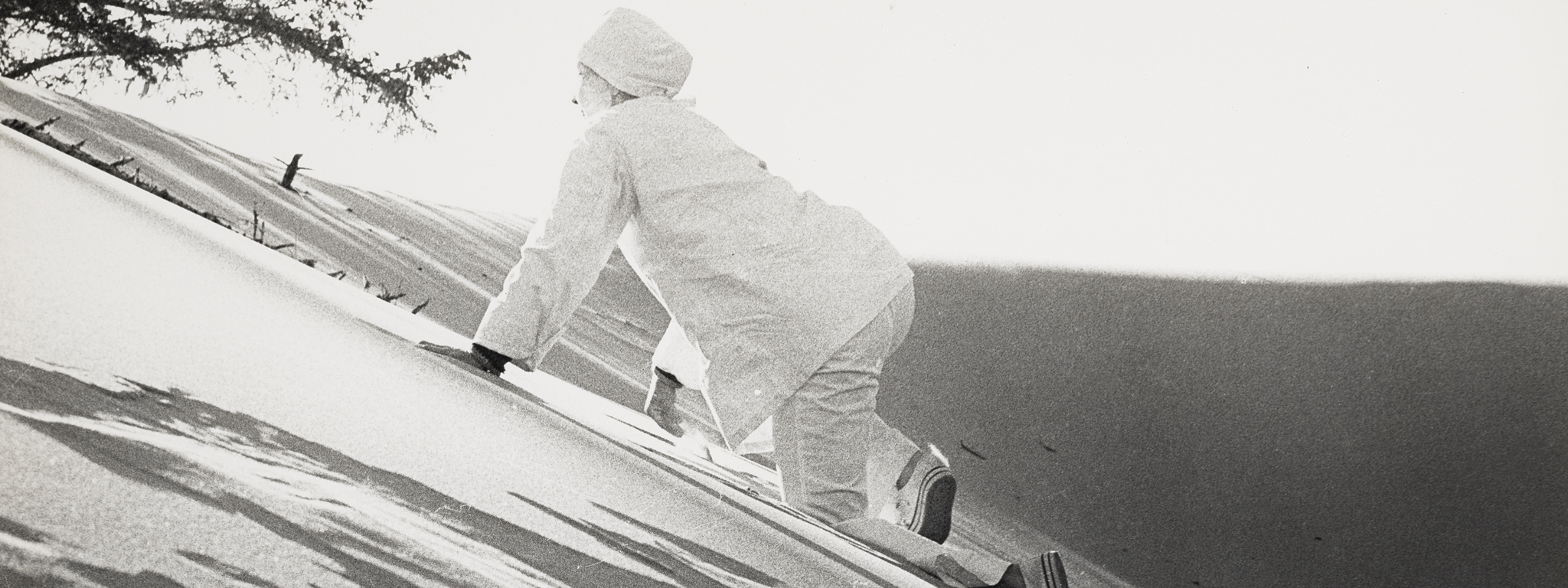 A black and white photograph shows a figure dressed in white with a white cap climbing a sand dune on hands and knees