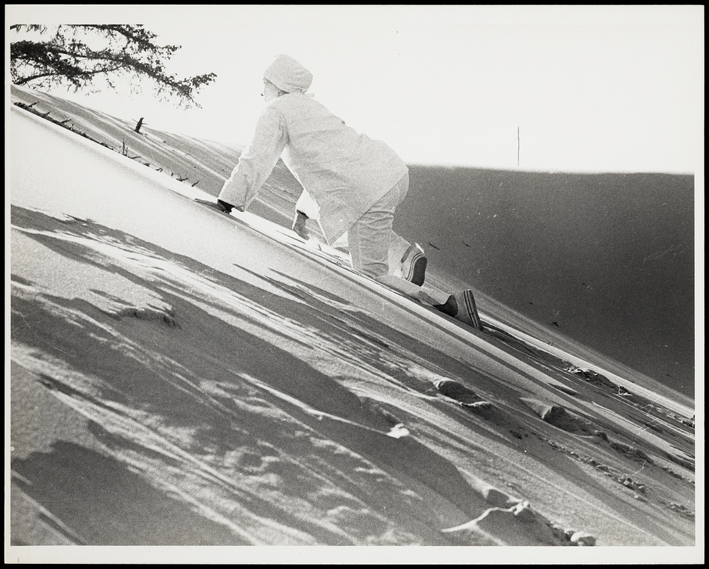 A black and white photograph shows a figure dressed in white with a white cap climbing a sand dune