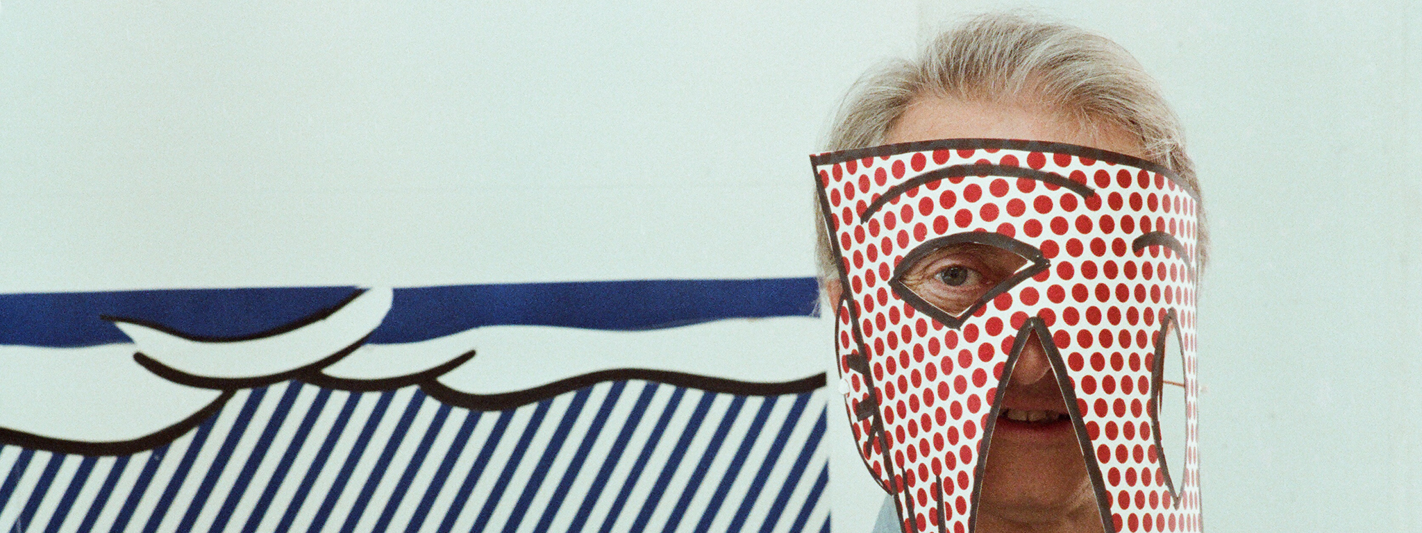 A man with graying hair in a denim shirt wears a paper mask covered in red dots. Behind him is a print made up of blue stripes and red dots