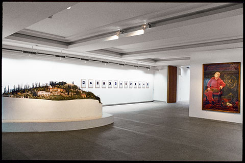 A color photograph documents a spacious gallery, occupied on the left by a large model of a verdant hillside estate that sits on a white base. A row of small, framed paintings lines the back wall, and the wall on the right side displays a larger painting of a blindfolded man dressed entirely in red 
